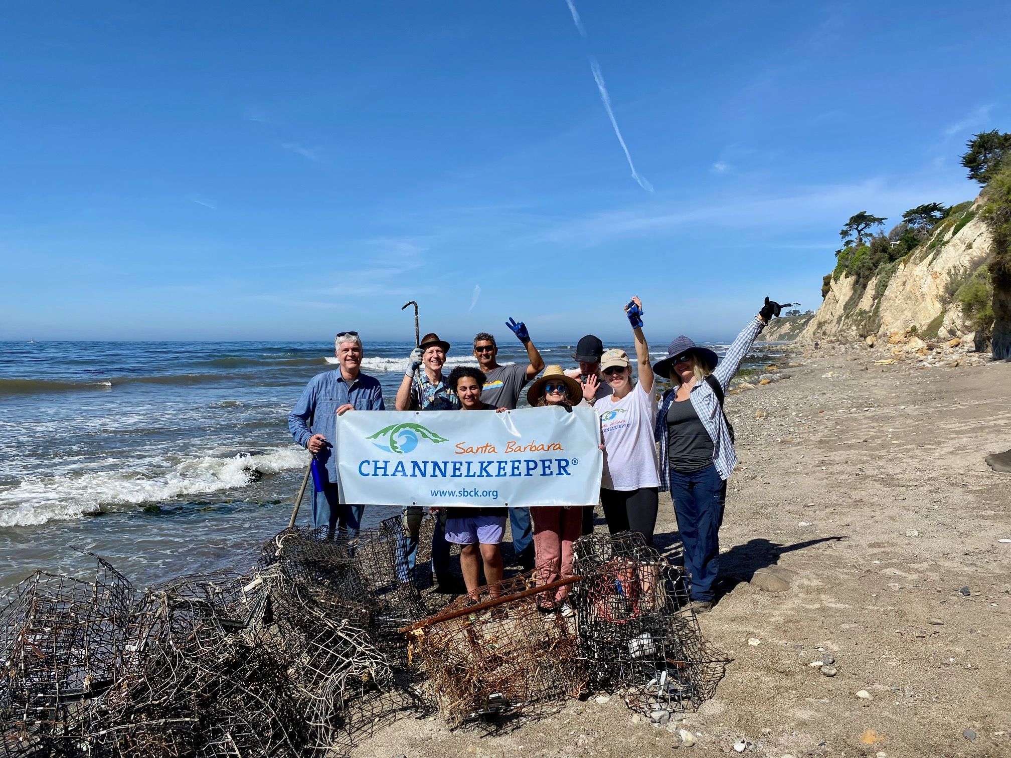 A group of volunteers on the beach with a pile of trash, waving and holding a banner reading Santa Barbara Channelkeeper